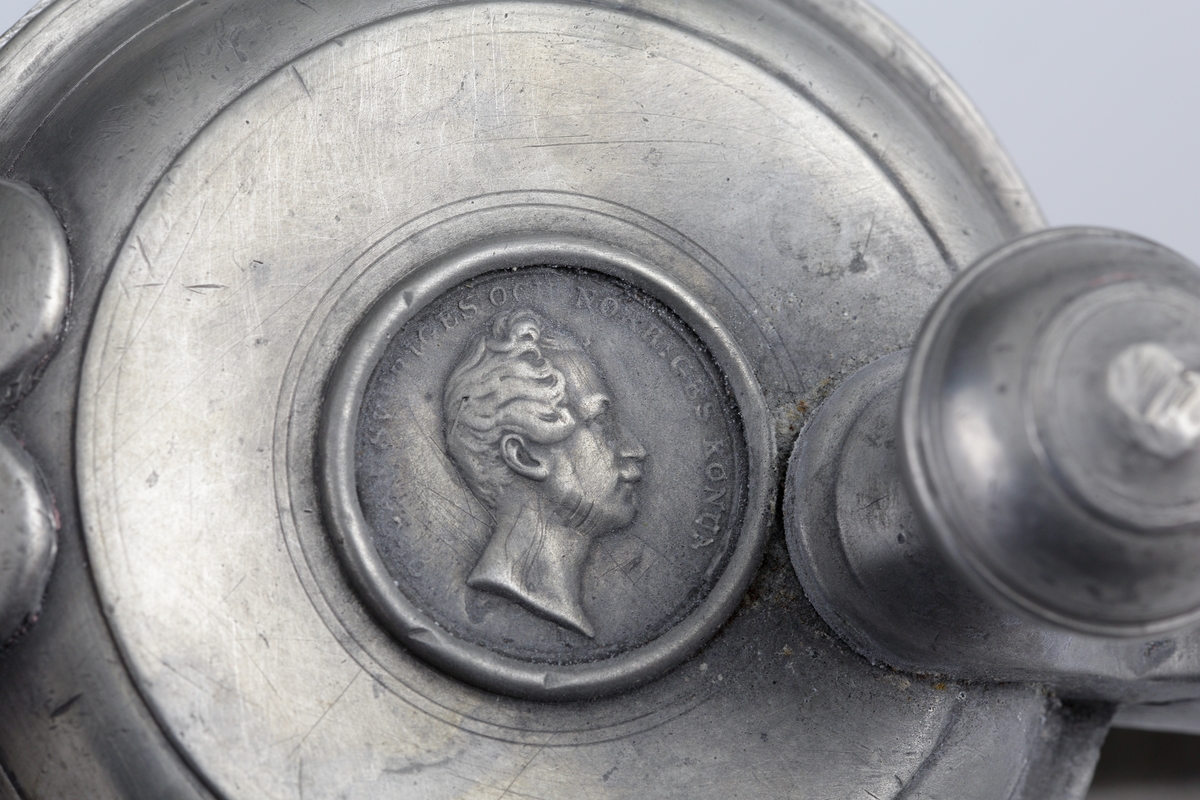 Stop av tenn.
Konisk modell. Svängd hänkel och tumgreppsknopp. Svängd, fasetterad snås. Fräcklad blomdekor på livet. På locket medaljong med Oscar I:s bild och inskriptionen: "Oscar, Sveriges och Norges konung". Stämplar på bottnens insida.

Inskrivet i huvudbok 1955.