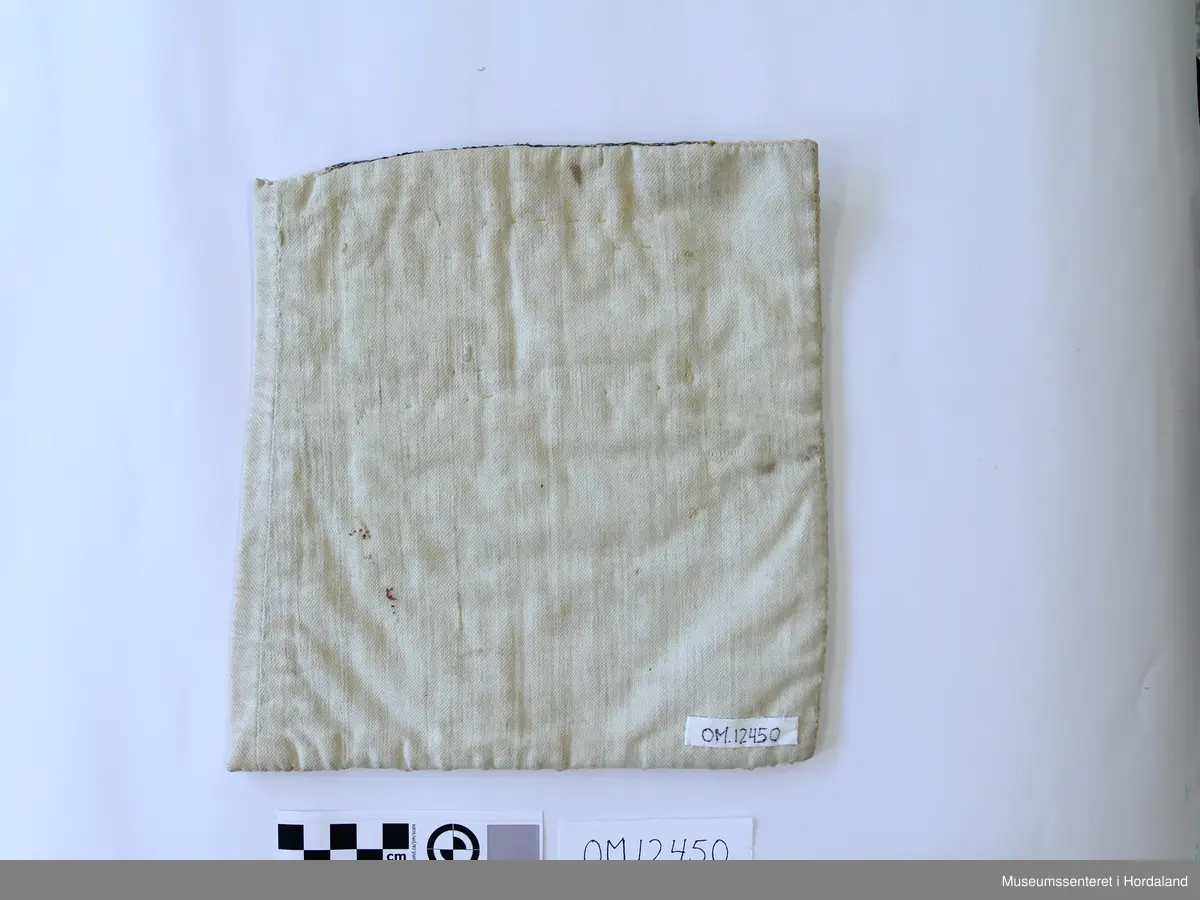 Bringeklut med ullbroderi i korssting i raud, grønt, gult og lilla. Sydd på stramei. Metallborder innimellom broderiet. Rosete silkevevnad oppe i blått,grønt og fleire raudlilla fargar. Eit silkeband nede i rosa, grøn og gul farge.  Montert på kvitt bomullsstoff.