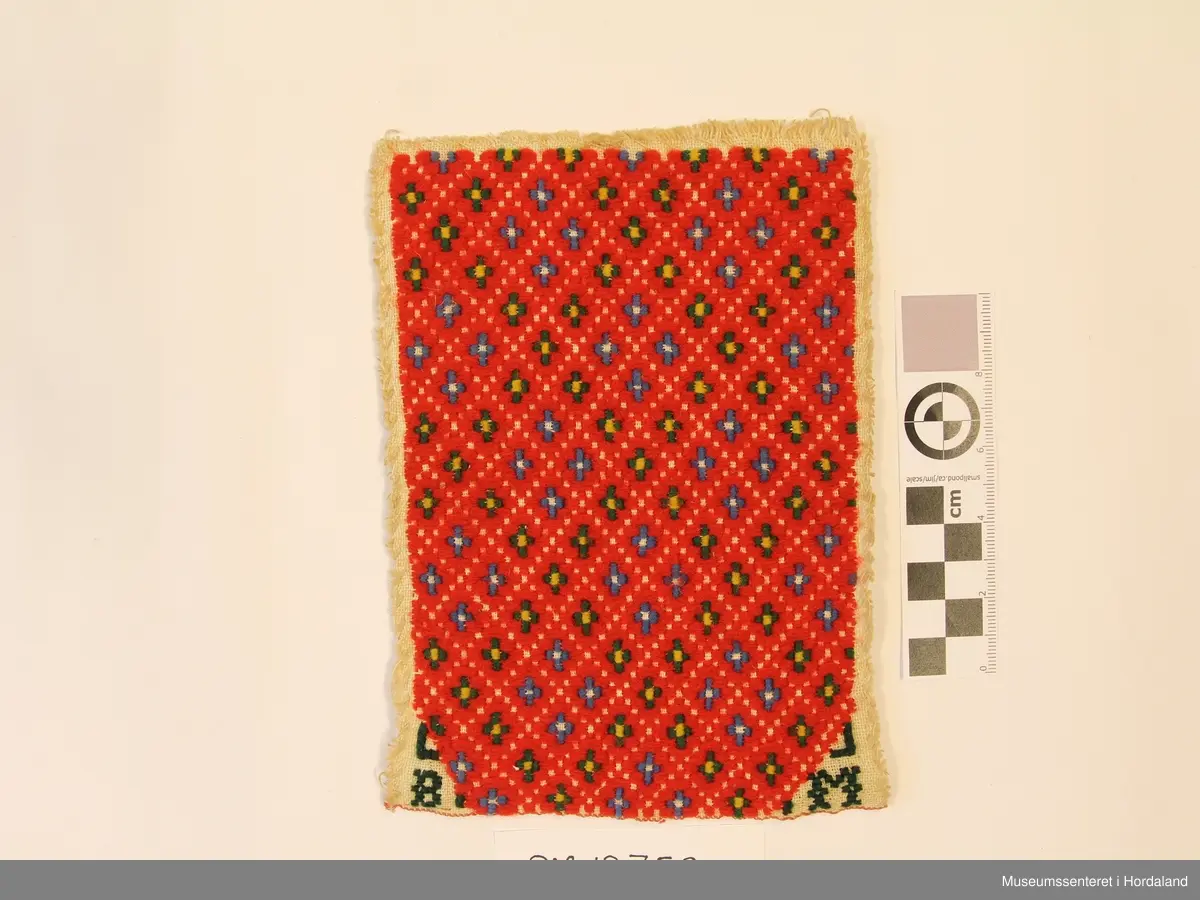 Broderi i plattsaum. Motivet er småruter. Nede i kvart hjørne er det brodert B i venstre hjørne og M i høgre hjørne. Brodert på kvitt linstoff.