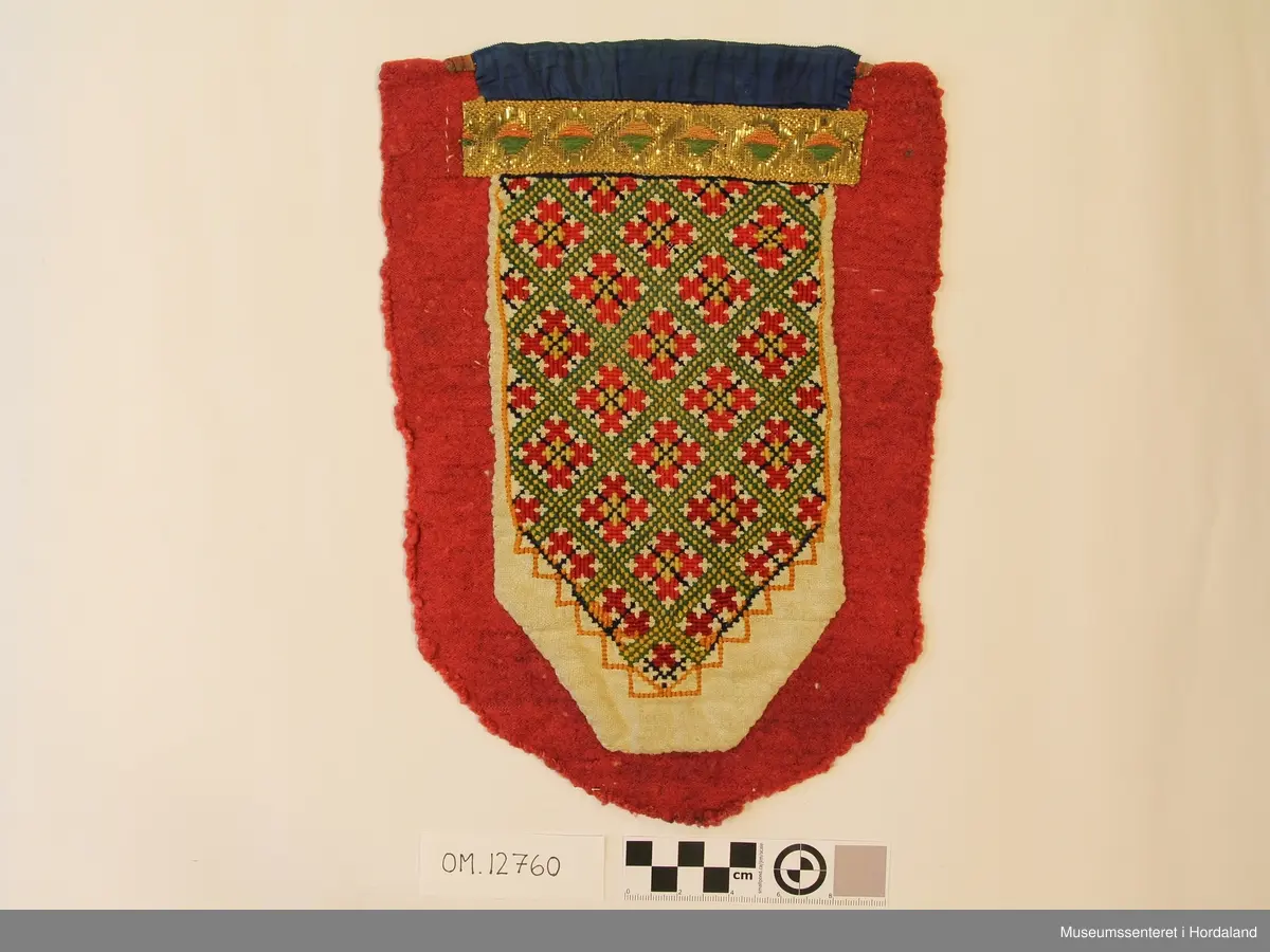 Broderi i kross-sting i ullgarn. Motivet er ein geometriske roser inndelt i ruter. Brodert på kvitt linstoff. Oppe er det eit forgylt sølvband, over der igjen eit blått sikeband. Alt er montert på eit raudt stykke ulltrikotasje. Oppe bak er det eit rutete bomullstoff.