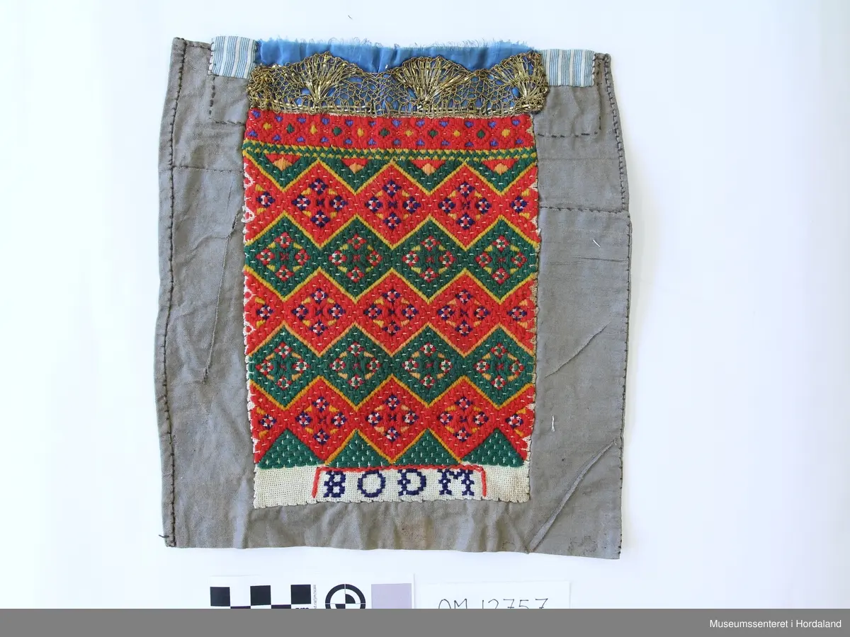 Brodert med ullgarn i plattsaum. Alt er handlaga. Hovudmotiv er små åttebladroser i border. Oppe er det påsydd eit blått silkeband med eit breitt gullband (i forgylt sølvtråd) utanpå silkebandet. Alt er montert på eit tynt, vokset bomullslerret. Oppe på baksida er det eit ekstra bomullstoff (blåstripet). Bokstavane BODM er broderte inn nede på bringeduken i kross-sting.