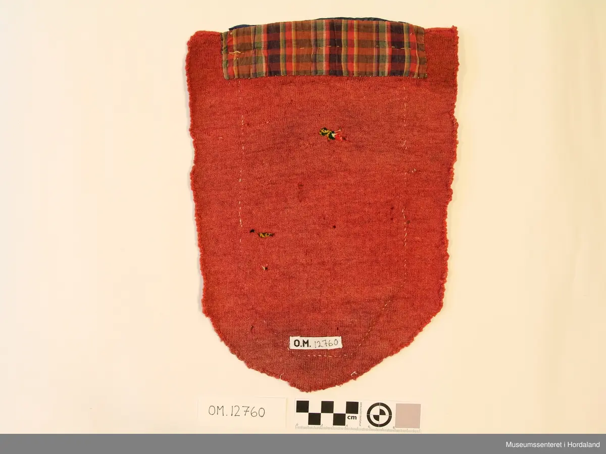 Broderi i kross-sting i ullgarn. Motivet er ein geometriske roser inndelt i ruter. Brodert på kvitt linstoff. Oppe er det eit forgylt sølvband, over der igjen eit blått sikeband. Alt er montert på eit raudt stykke ulltrikotasje. Oppe bak er det eit rutete bomullstoff.