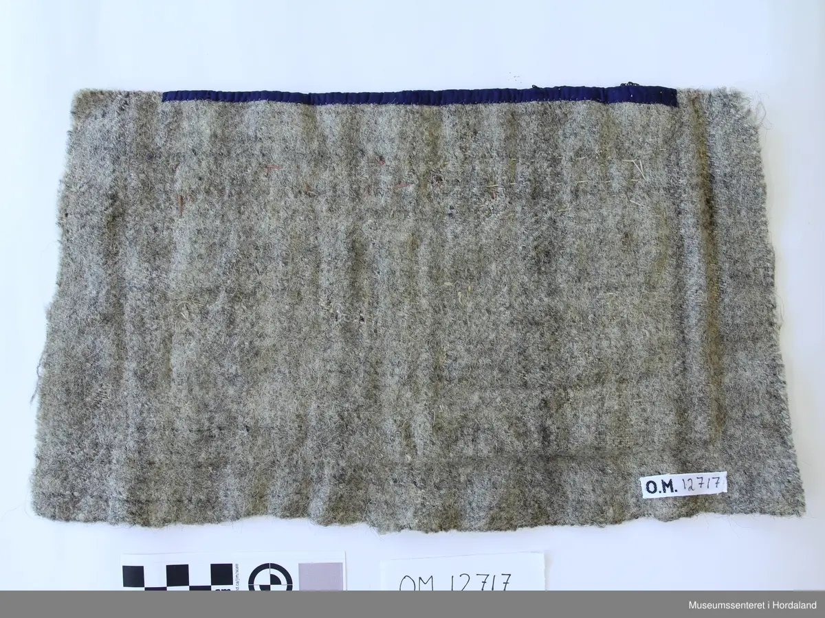 Rektangulær bringeduk med 2 rader perlenett montert på raudt stoff - annakvar perlerad og metallband. Både sølv og gull metallband. Kronekvarde oppe montert på blått band.  Bredda er større enn høgda. Bakstykke er eit vove ullstoff i kypert.