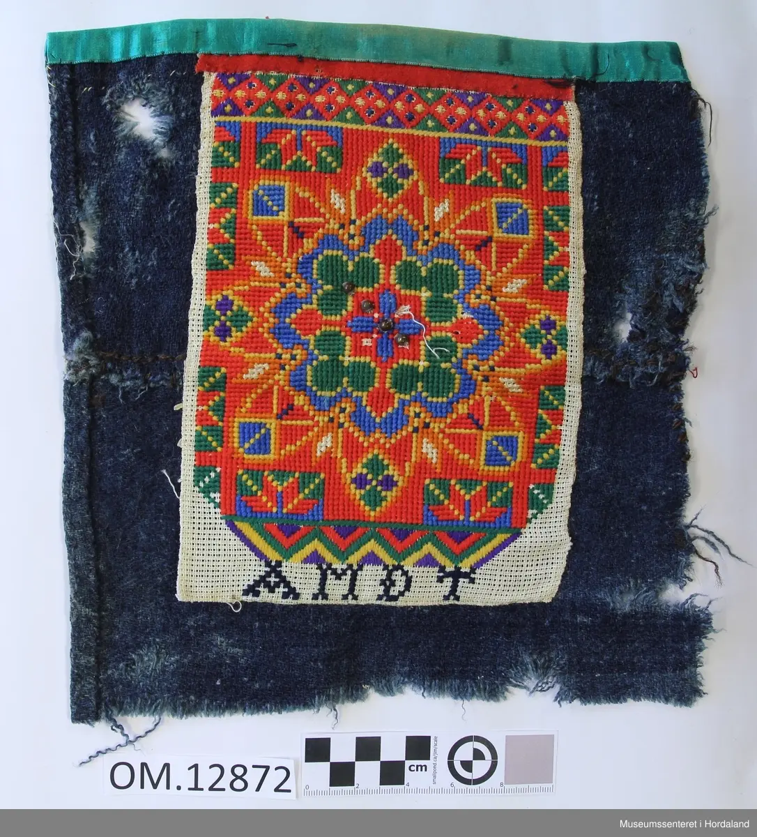 Rektangulær bringeduk på kvadratisk blått, kypertvevd bakstykke i ull. Broderiet er sydd på stramei. Blomstermotiv i midten og diverse geometriske figurer. Bord oppe og nede. Plattsaum og enkel kross-sting i fleire retningar. fire store glasperler inne i blomsten- Brodert i kross-sting under broderiet A M D T. Turkis band øvst oppe på kanten, eit raudt ullstoff imellom den turkise kanten og broderiet. Holete. Bakstykket er sydd saman av to deler og lappa. Kant lagt inn på eine sida.