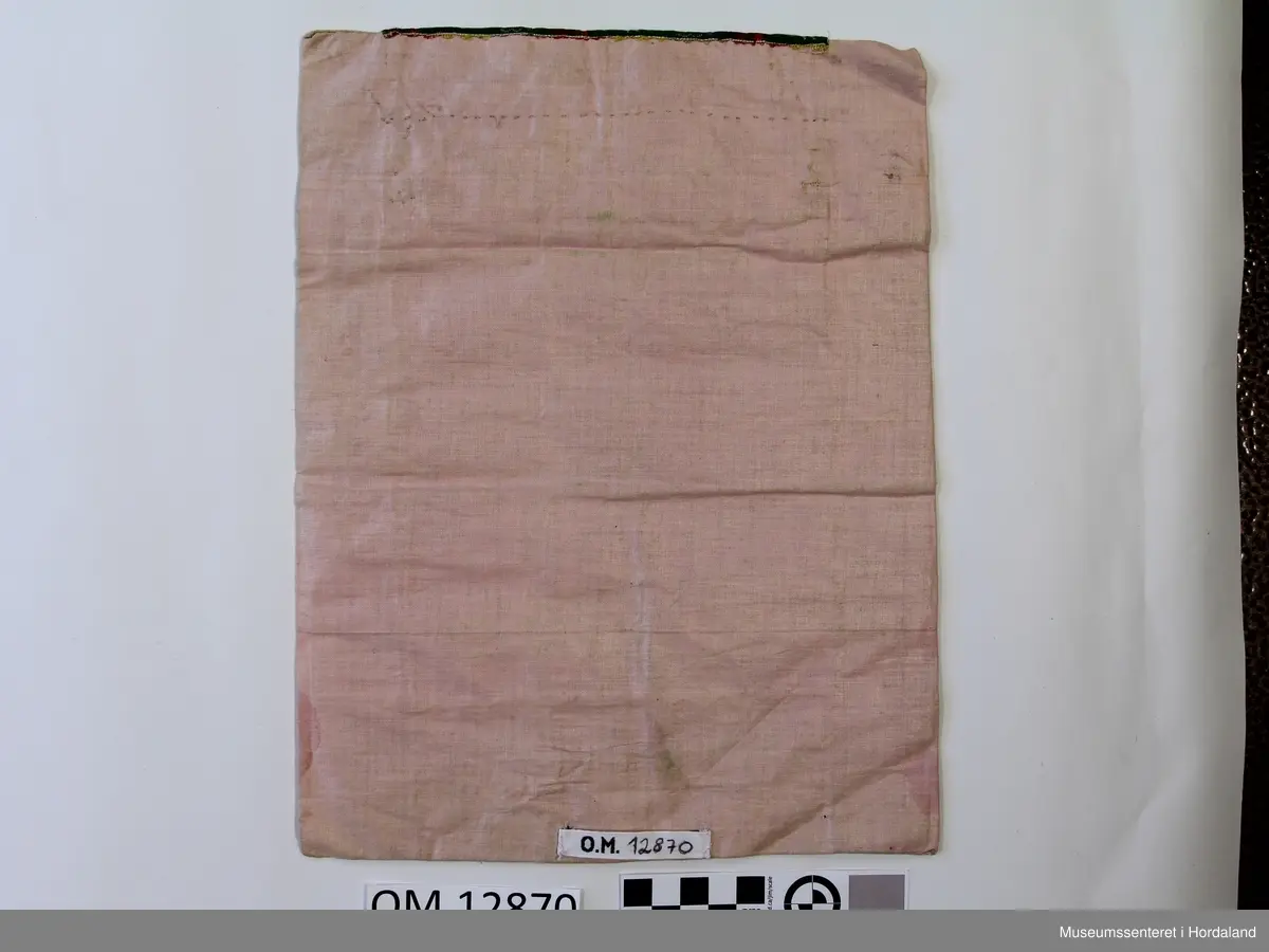 Rektangulær bringeduk med raudt ullstykke framme uten brodering. Oppe er det ein kant med gullfrga metallbord oppå eit brokadeband, metallborden går og eit stykke ned på det raude stoffet. Rosa, blankt fõrstoff til brei kanting og bakstykke. Alt er handsauma. Eit stykke papir el l inni til forsterking. Litt felkkete på forstoffet.