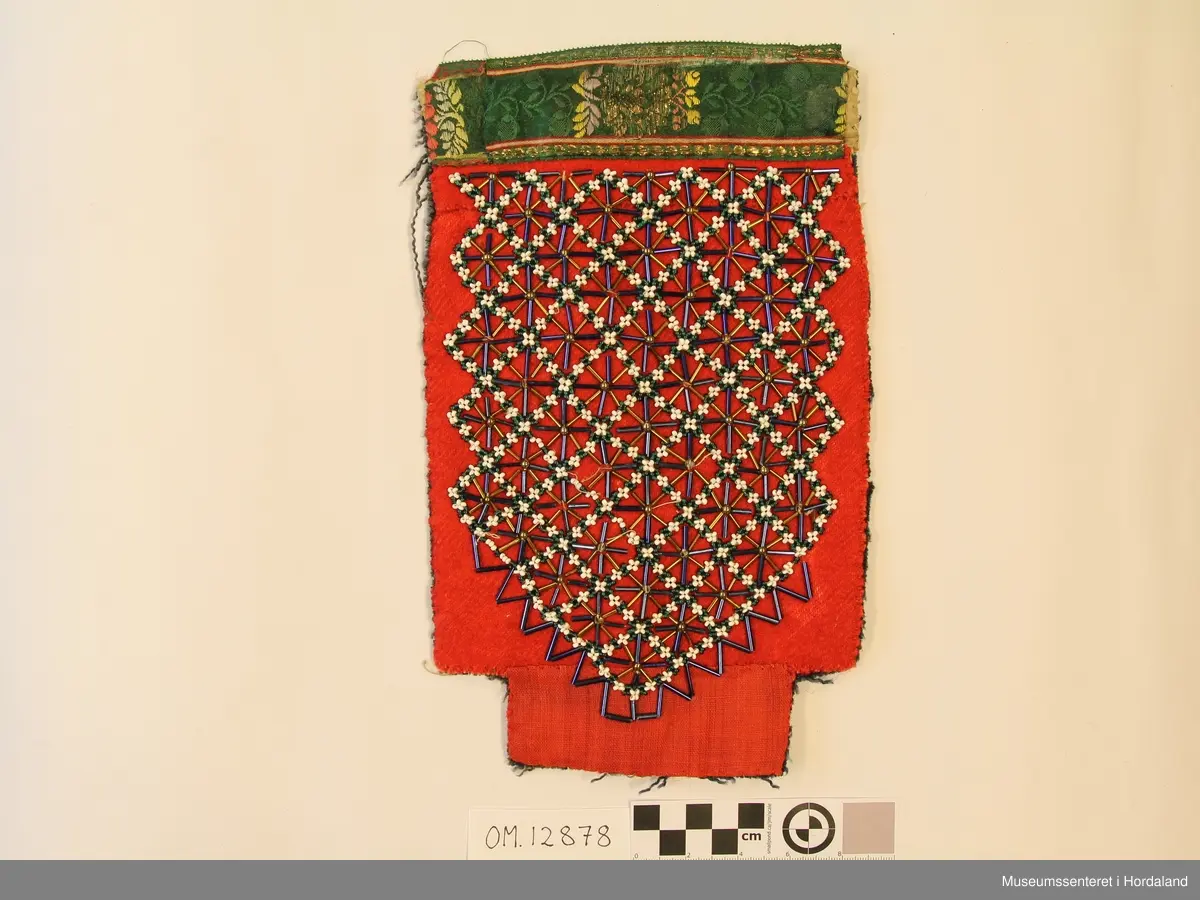 Rektangulær brineduk med perlebroderi/perlenett på raudt ullstoff. Skjøtt på eit lite stykke nede i eit tynnare ullstoff.  Silkeband oppe til kant. Bakstykke av blå ullkypert, fillete. Bomullstoff i øvre kanten bak bandet til forsterking.