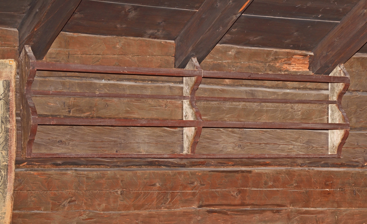 Kärlhylla av furu, brunmålad, långsmal med två hyllplan, profilerade gavlar samt mittpost. Två längsgående lister håller kärlen på plats.