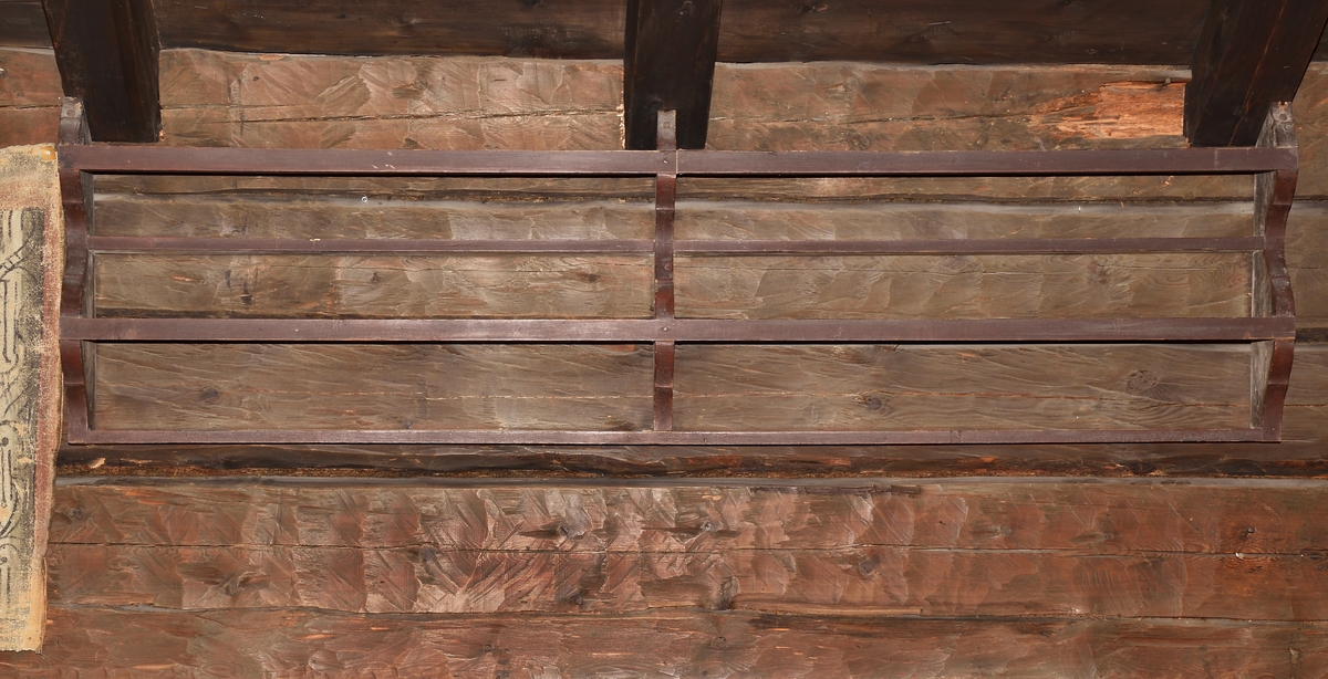 Kärlhylla av furu, brunmålad, långsmal med två hyllplan, profilerade gavlar samt mittpost. Två längsgående lister håller kärlen på plats.