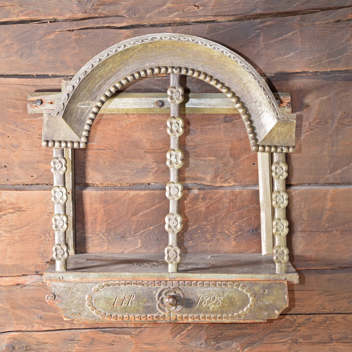 Tavlett av grönmålad furu med låda. Bågformigt krön med skuren pärlstav, uppburet av tre pelare med utskurna blommor. Låda med blomma och svarvad dragknopp. Lådfrontens hörn i form av enkel volutformation, högra hörnet saknas dock.