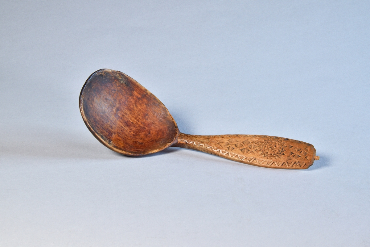 Sked av lövträ med riklig karvsnittsornamentik på skaftet. Djupt blad, böjt mot skaftet. Spår av grön bemålning på bladet.