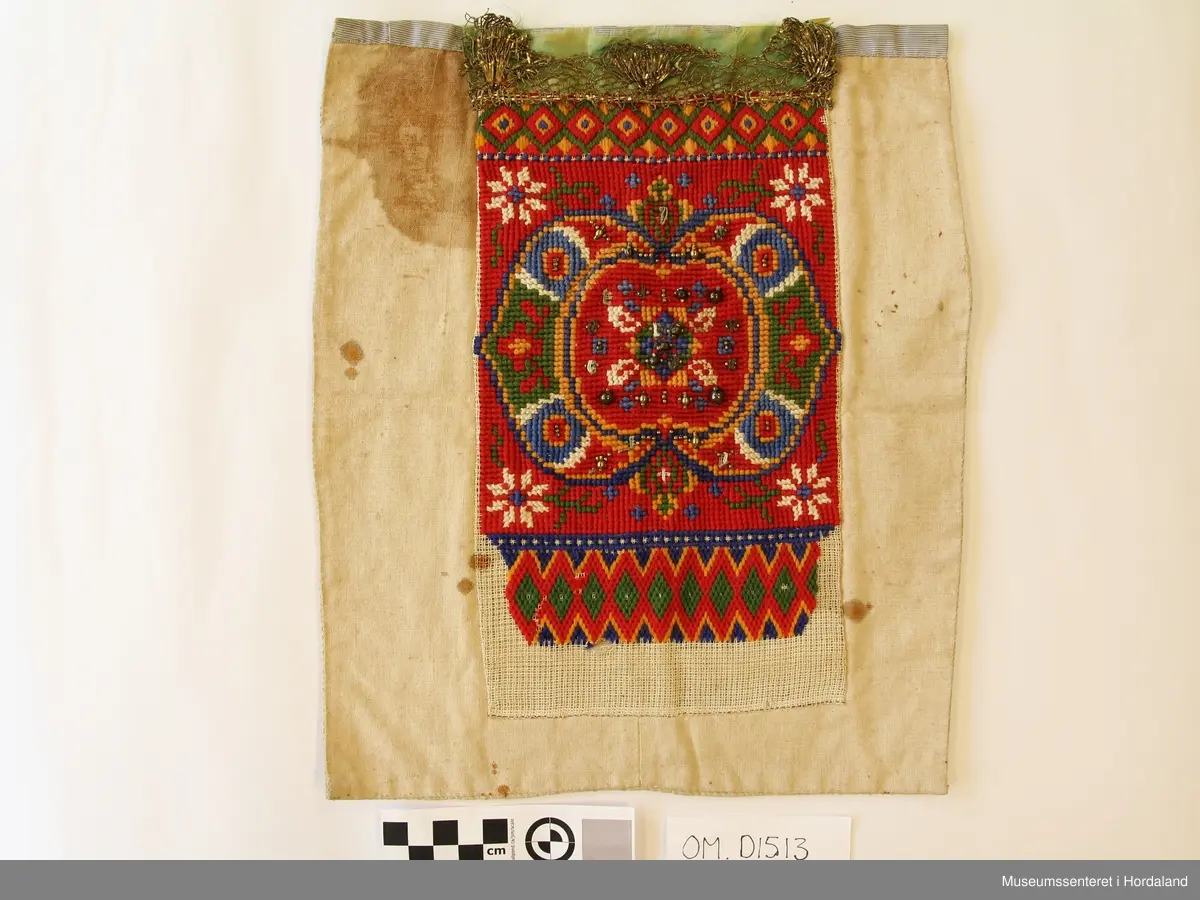 Bringeklut brodert på stramei med kross-sting og dragesaum. Raud med blått, gult, grønt og gull. Festa på eit kvitt stoff. Stoffet har ein stor flekk og nokre små. Oppe på baksida er montert på eit stripete stoff som går eit lite stykke ned på framsida. Grønt silkeband oppe med gullfarga metallkronekvarde sydd utanpå. Store og små perler sydd på broderimønsteret, nokre av desse er knuste.