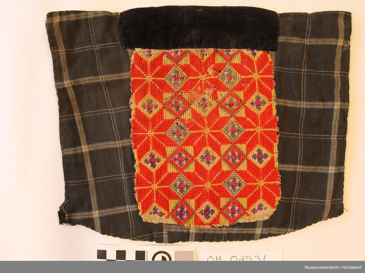 Rektangulær brystduk som er brodert i kross-sting (halve og heile). Åtteblad-rose som hovudmotiv og ruter. Broderiet er litt slitt. Svart fløyelsband oppe. Broderiet er sydd på linstoff. Bakstykket og sidestykke er i bomull - rutete mønster.