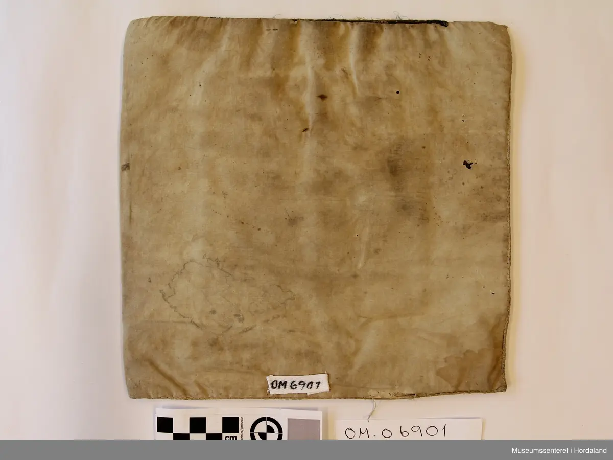 Rektangulær bringeduk sydd på kvadratisk bomullstoff. Broderiet er i halve og heile kross-sting sydd på linstoff. Raud hovudfarge. Initialane I O D M er sydd nederst på broderiet. Silkeband øvst med ei smal metallbord nedom. Silkebandet, metallborda og broderioet er veldig slitt.