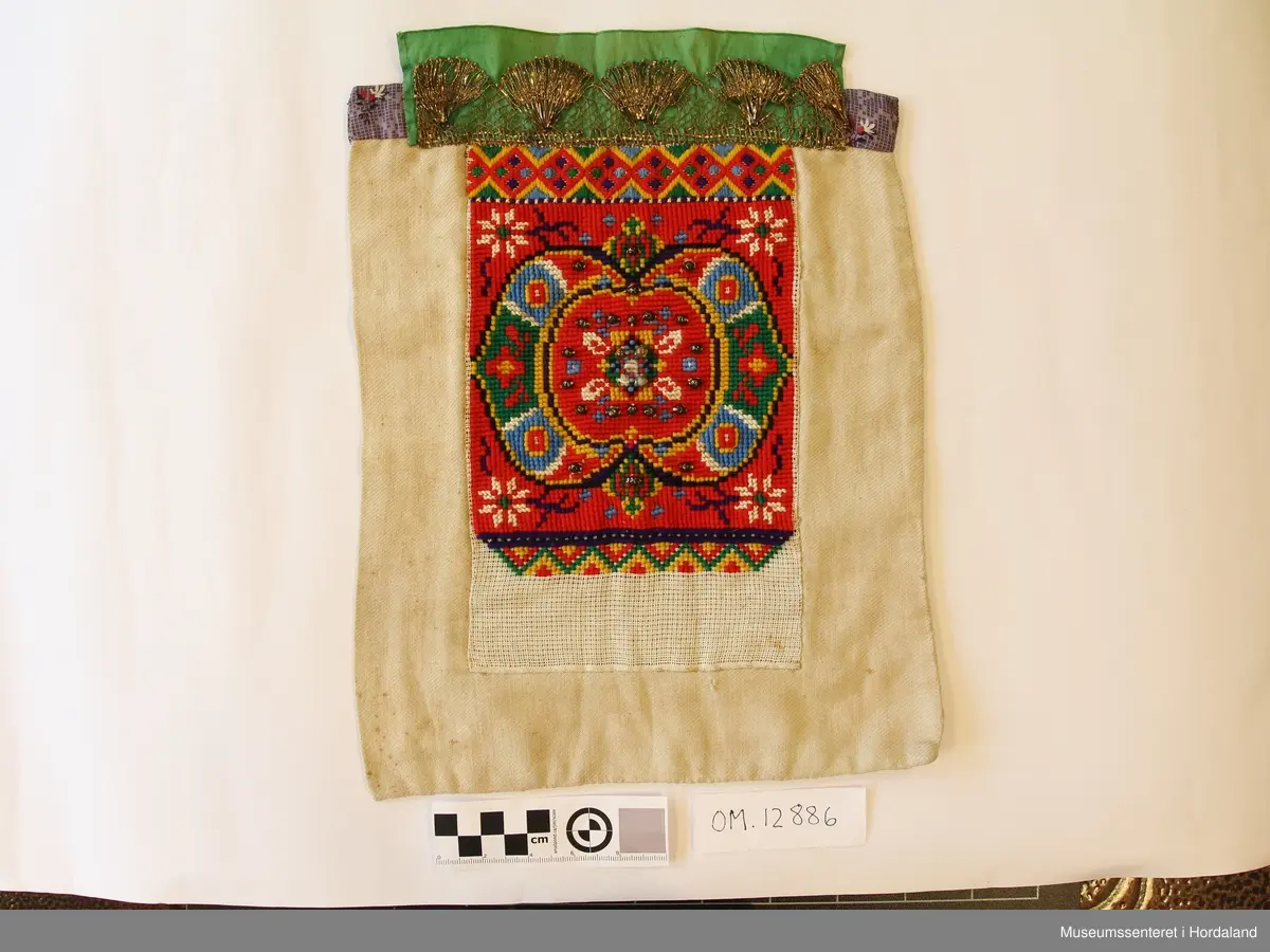 Kvadratisk bringeduk med motiv i kross-sting - hovudmotivet er dekorert med nokre store perler (to knuste). Metallkvarde oppe på eit grønt silkeband. Lilla band bakom oppe. bakstykke på eit stoff som er naturfarga.