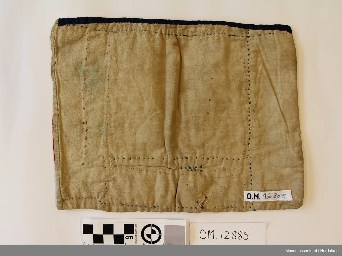 Rektangulær brystduk med raude stoff på sidene og eit bomullstoff på baksida. Blått band oppe. Firkanta broderi i kross-sting, med opne ruter. Åttebladrose og firkantmønster.