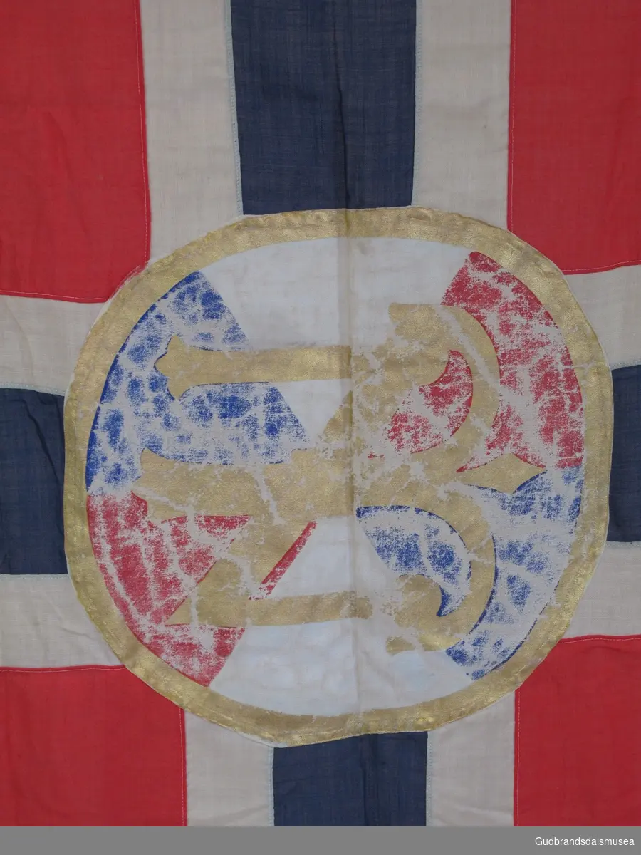 Norsk flagg med Den norske turistforenings logo påmalt i midten. Innsydd line øverst med metallhengsel i ene enden. I bruk på¨Sandhaug turisthytte frem til 1948.
