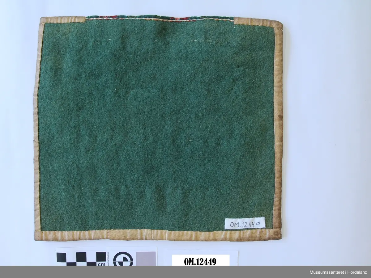 Bringeklut med ullbroderi i smøyg i fargane raudt, grønt og gult. Smale metallborder innimellom broderiet. Rosete silkeband oppe i grønt, gult og to raud/lilla fargar. montert på kvitt bomullsstoff som no er veldig gulna og flekkete. Bakstykket er i grønt vadmål.