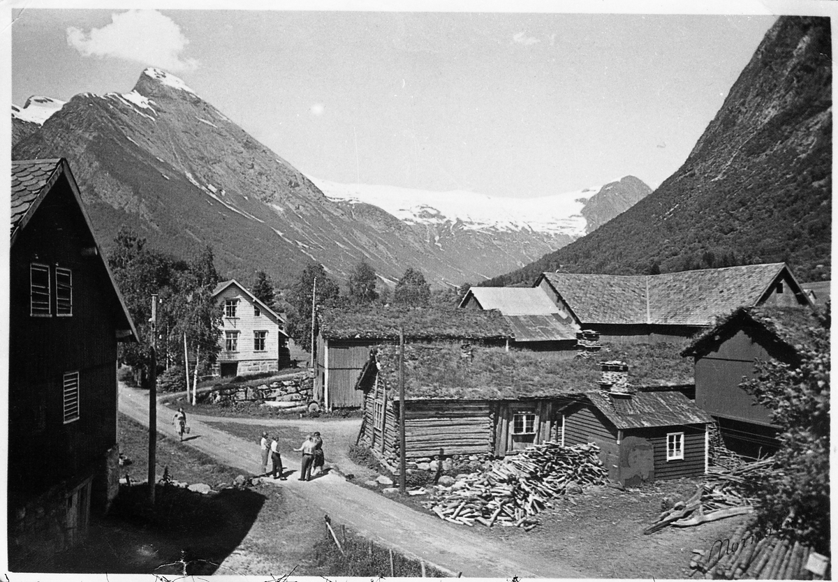 Avfotografert bilde fra tunet på en fjellgard. På tunet er noen uidentifiserte personer. I bakgrunnen fjell som delvis er snødekte. Hverken personer eller sted er identifisert.