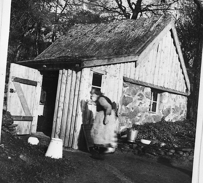Josefina "Skrape-Fina" Karlsson vid sin backstuga i Dagsås på 1950-talet. Det förefaller som att såväl gaveln som boden fått ny panel nyligen.