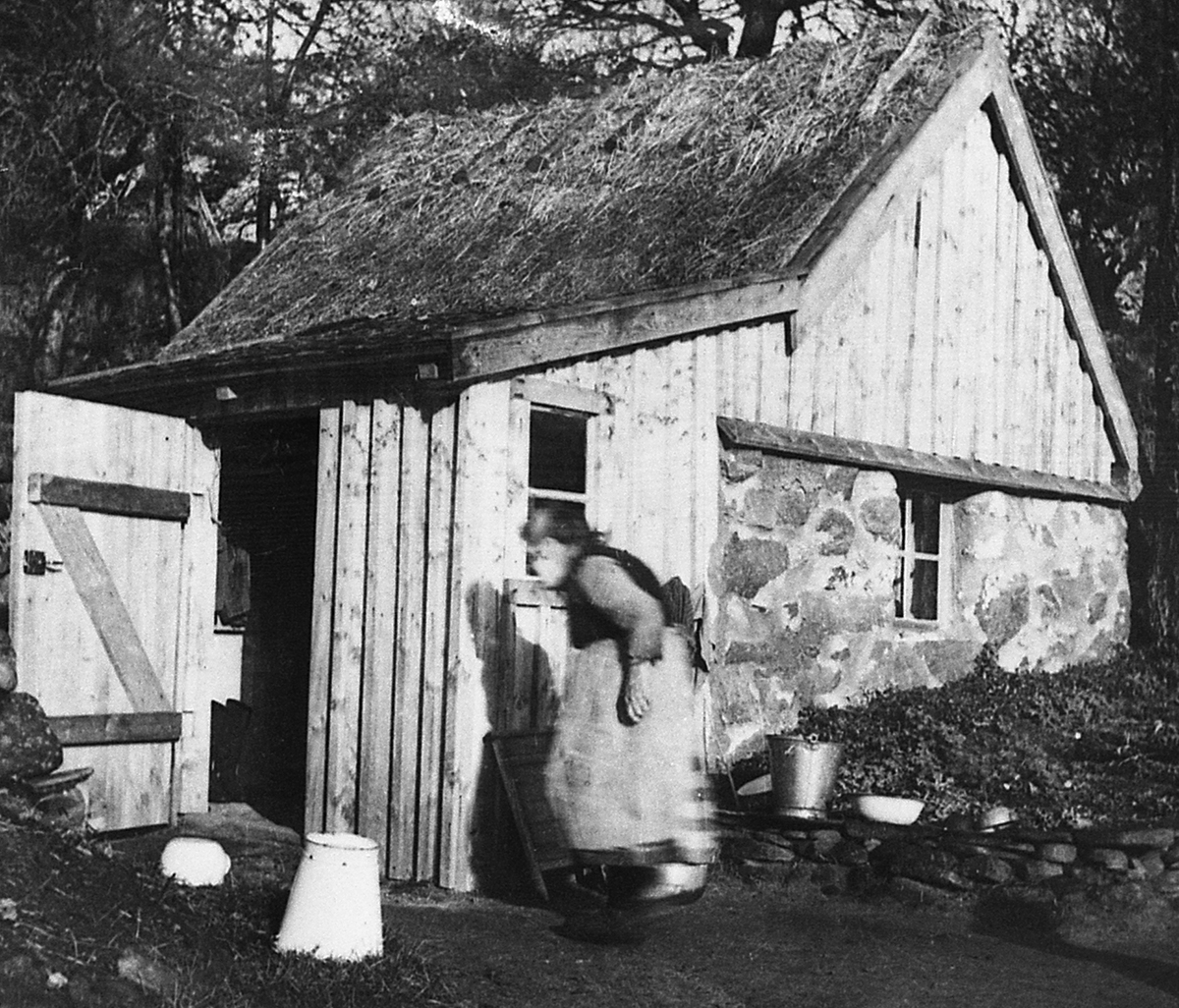 Josefina "Skrape-Fina" Karlsson vid sin backstuga i Dagsås på 1950-talet. Det förefaller som att såväl gaveln som boden fått ny panel nyligen.
