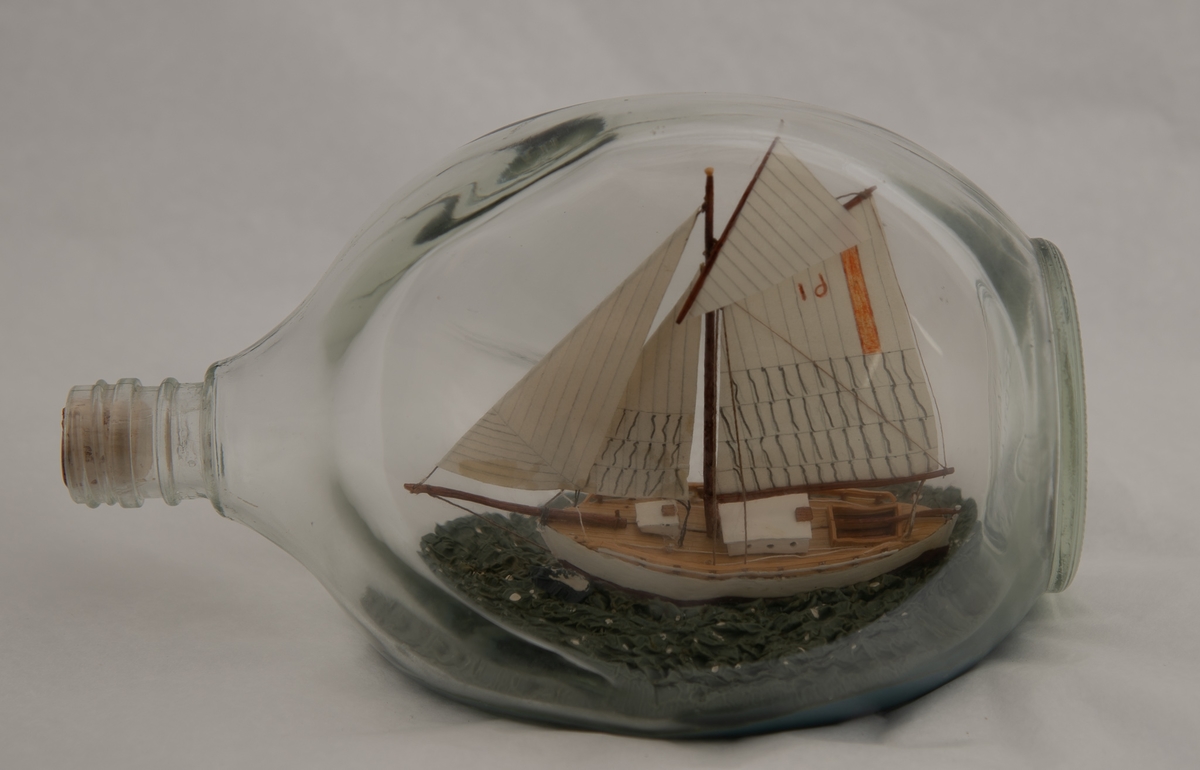 Flaskeskute: "Krabben" , losbåt nr. l (Colin Archer)  Flaske i klart glass med trekantet form.  Hvitmalt skrog , brun under vannlinjen. Seil med oransje stripe og tallet 61 i oransje. Blågrønn sjø.