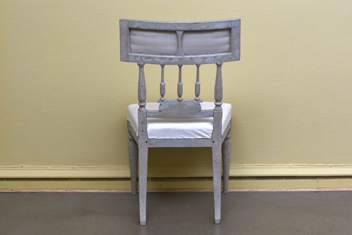 Gråvit stol i gustaviansk stil av björk på runda, avsmalnande ben. Benens övre del är dekorerade med bladkrans. Den främre sargen är något svängd och dekorerad med en äggstavslist. Den rektangulära ryggbrickan bärs upp av totalt fem, tre mindre och två större, ståndare. Ryggbrickans ramvirke är dekorerad med äggstavslist. Stolen är provisoriskt klädd på sits och i rygg med ett vitt bomullstyg.