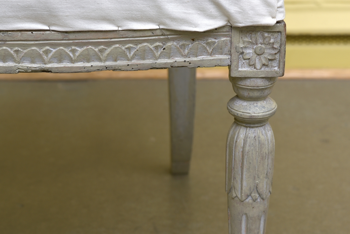 Gråvit stol i gustaviansk stil av björk på runda, avsmalnande ben. Benens övre del är dekorerade med bladkrans. Den främre sargen är något svängd och dekorerad med en äggstavslist. Den rektangulära ryggbrickan bärs upp av totalt fem, tre mindre och två större, ståndare. Ryggbrickans ramvirke är dekorerad med äggstavslist. Stolen är provisoriskt klädd på sits och i rygg med ett vitt bomullstyg.