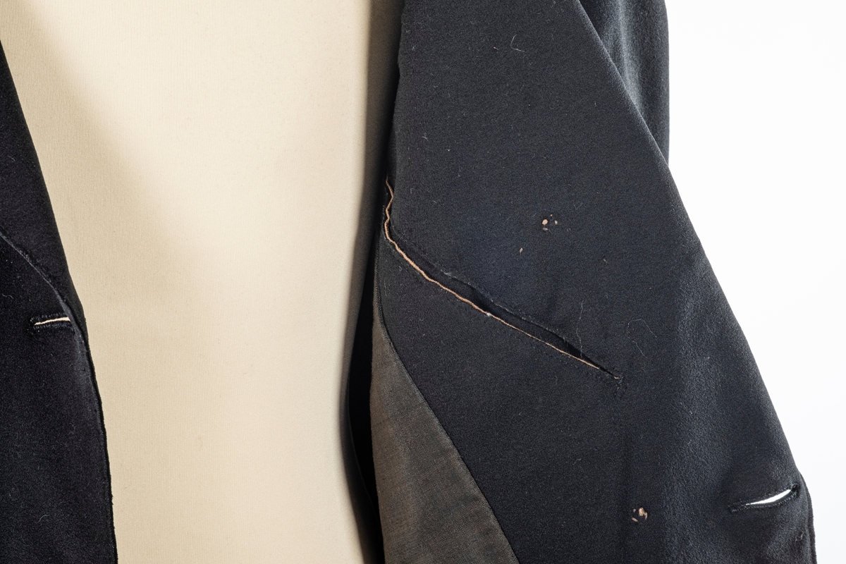 Dobbelt spent frakk som kneppes med fire knapper på hver side. De øverste kan ikke kneppes. Knappene er kledd med silke med fløyelsornament i midten. Hempe i nakkelinning. Det er splitt med legg fra livet og ned, med en knapp over splitten på hver side. Det er også to knapper lenger ned i splitten, og en knapp på hver mansjett. Frakken er foret.