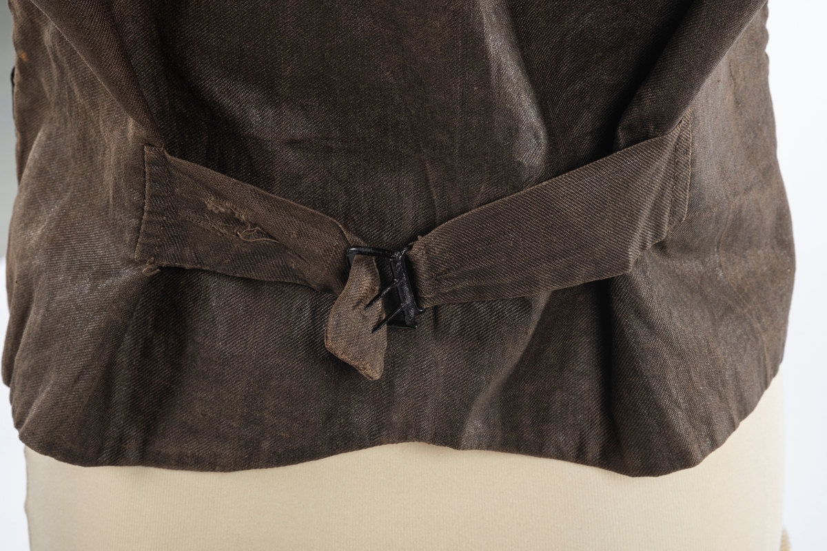 Vest med forstykke i silke og bakstykke i bomull. Den er foret med lin. Det er dobbelspent knepping og spensel i rygg. Knappene er trukket med silke.