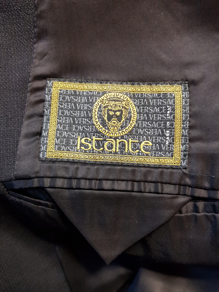 Hoftelang, dobbeltspent, med 6 metallknapper, med Versace-husets "Istante"-logo - Zevs hode - i relieff. Lange slag, og kun de nederste 4 knapper skal kneppes. Lapellene (slagene) er spisse og lengre enn kragen. Ett ekstra knapphull for innvendig knapping, men knappen på innsiden av venstre forstykke mangler. To-søms ermer med falsk splitt, lukket knapphull og pynteknapp. Horisontal lomme med belegg på hver hofte og stikklomme med belegg på venstre bryst. Innvendig, passepoilert lomme på hvert bryst, den høyre lukket med klaff og usynlig knapp. Søm midt bak og splitt nederst i hver panelsøm bak. Forstykkene er skåret rettvinklet i nederkant midt foran. Helfôret med svart fôrsilke. Kragestanden og kragen er kledd med filtaktig materiale på baksiden og jakken har puter i skuldrene.