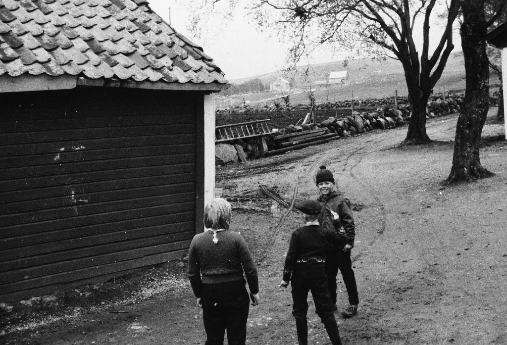 Kjellaug Kverneland og Arne Kverneland og Jørn Lemvig leikar med pilkasting på løeveggen. I bakgrunnen husa til Thu, Stangeland og Helgeland på Veidene.