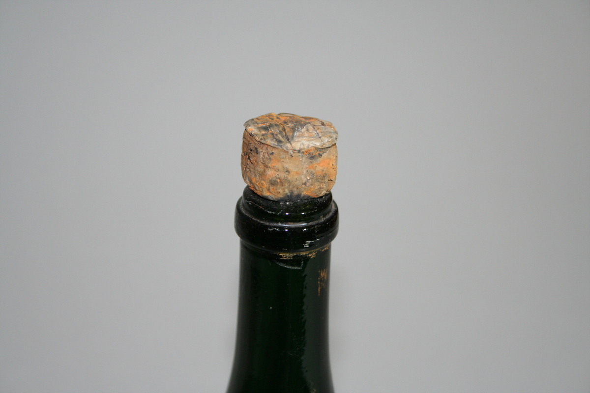 Tom flaske i farget glass og med opphøyet bunn. Korken sitter fortsatt på og har en "champagnekork" fasong. Et papir sitter tapet fast med informasjon om giver