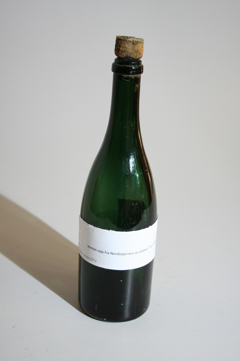 Tom flaske i farget glass og med opphøyet bunn. Korken sitter fortsatt på og har en "champagnekork" fasong. Et papir sitter tapet fast med informasjon om giver