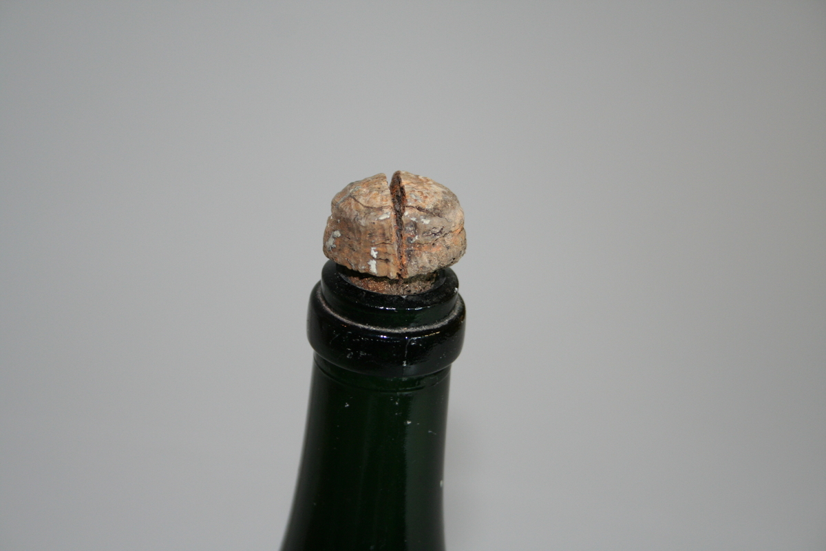 Tom flaske i farget glass og med opphøyet bunn. Korken sitter fortsatt på og har en "champagnekork" fasong. Korken har en sprekk i tioppen. Et papir sitter tapet fast med informasjon om giver