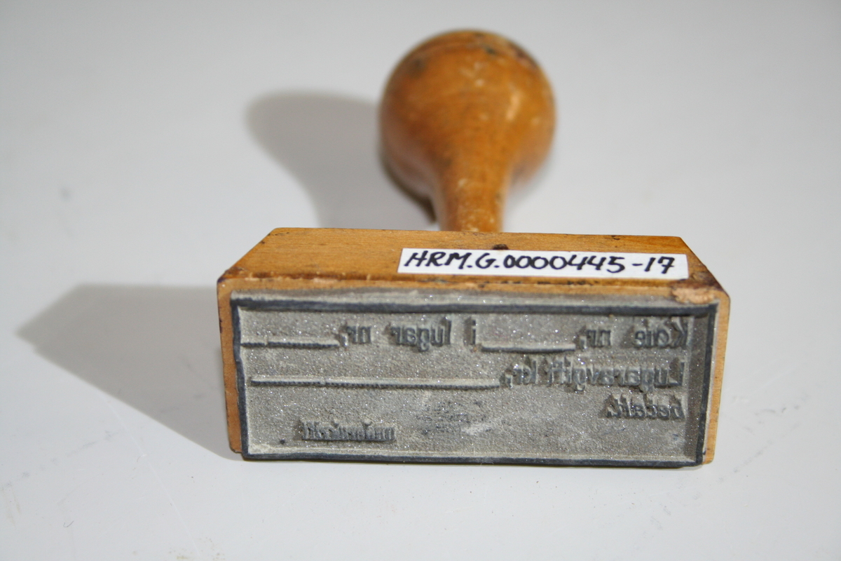 Stempel i tre med retangulær stempelflate. Dreid skaft. Et ovalt lite metallskilt på siden med produsentlogo. Lakkert
