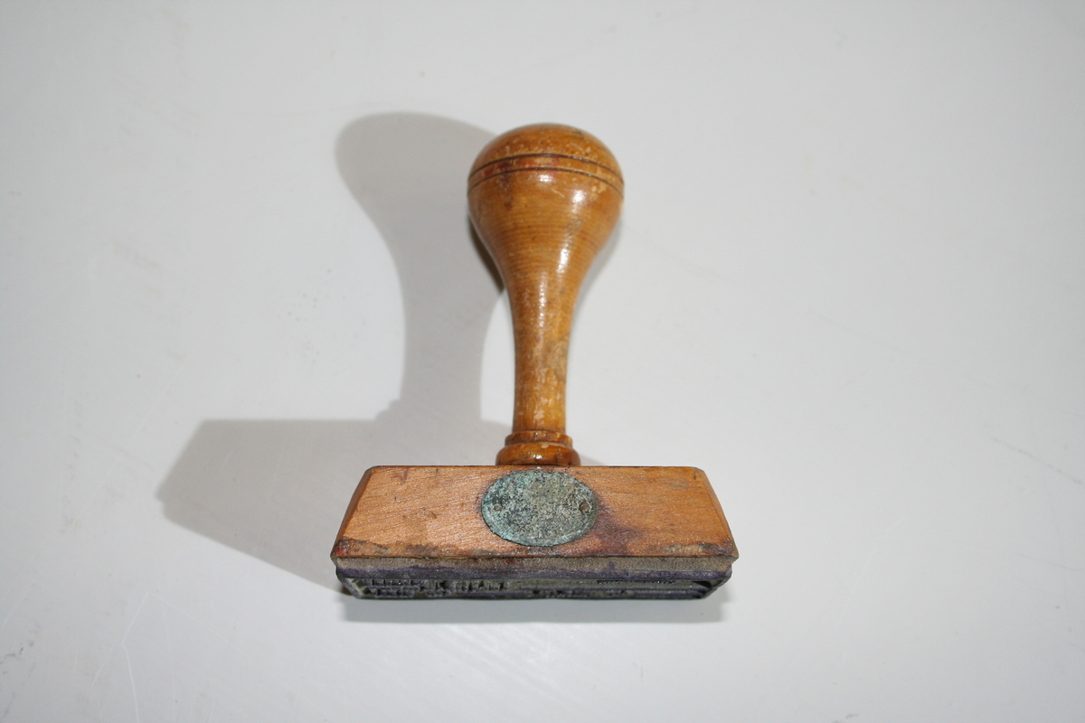 Stempel i tre med retangulær stempelflate. Dreid skaft. Et ovalt lite metallskilt på siden med produsentlogo. Lakkert