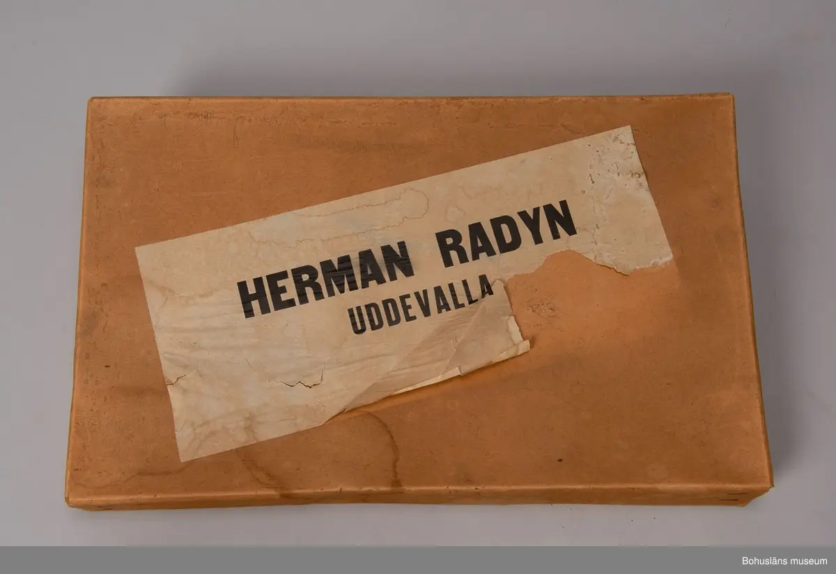 Låda av brun papp. På locket påklistrad,delvis bortsliten etikett med text: "Herman Radyn Uddevalla". Lådan innehåller: 19 st. julgransljushållare, glitter i silver, glitterband i guld, 7 st. julgranskarameller av papper, 1 toppstjärna, 12 st. hjärtan av papper i olika färger, 6 st. små glasbollar i sytråd, svenska flaggor i tråd, 12 st. svenska flaggor med järntråd,