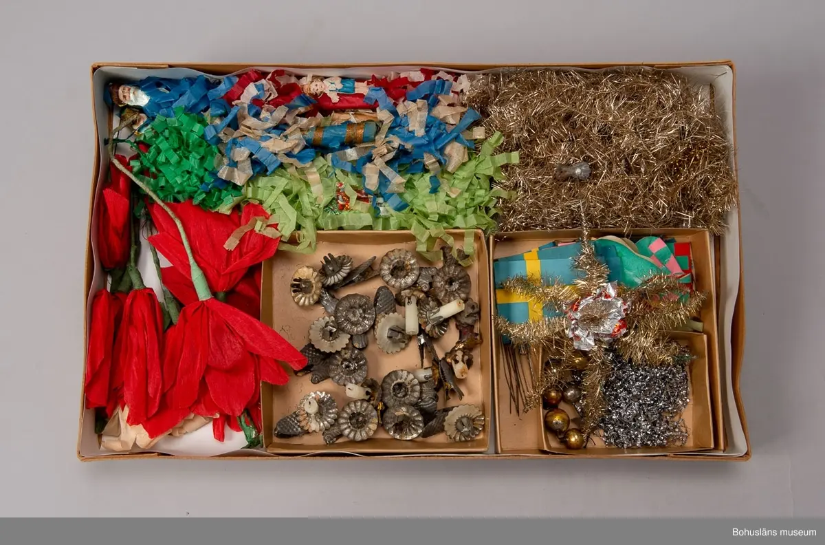 Låda av brun papp. På locket påklistrad,delvis bortsliten etikett med text: "Herman Radyn Uddevalla". Lådan innehåller: 19 st. julgransljushållare, glitter i silver, glitterband i guld, 7 st. julgranskarameller av papper, 1 toppstjärna, 12 st. hjärtan av papper i olika färger, 6 st. små glasbollar i sytråd, svenska flaggor i tråd, 12 st. svenska flaggor med järntråd,