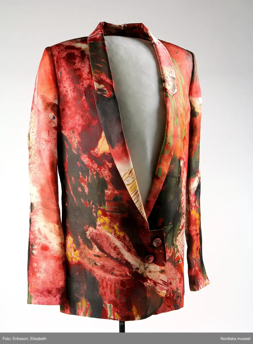 Blazer för kvinna, "Woman's Blazer", maskinsydd av mönstrat, tryckt sidentyg i rak modell med smala slag. Två diagonalt placerade klädda knappar framtill, fickor infällda i sidosömmarmarna. Rakt skuren nedre kant, rundad i mötet framtill, sprund nedtill i ryggen. Det tryckta sidentyget i röda nyanser har även inslag av brunt, grönt, gult och vitt. Innerfoder av  crèmefärgat sidentyg, smal beige etikett av läder med text: Whyred, fastsydd på insidan vid nacken (lossnat på ena sidan). I brösthöjd en innerficka på vardera sida, knäppt med klädd knapp. Under vänstra fickan en beige sidenetikett med text: WHYRED, samt fastsydda i etikettens nedre söm, vita sidenband med tryckt text: 36. Made in China.
Lisa Eide 2015-03-26, Helena Lindroth 2019-01-21
