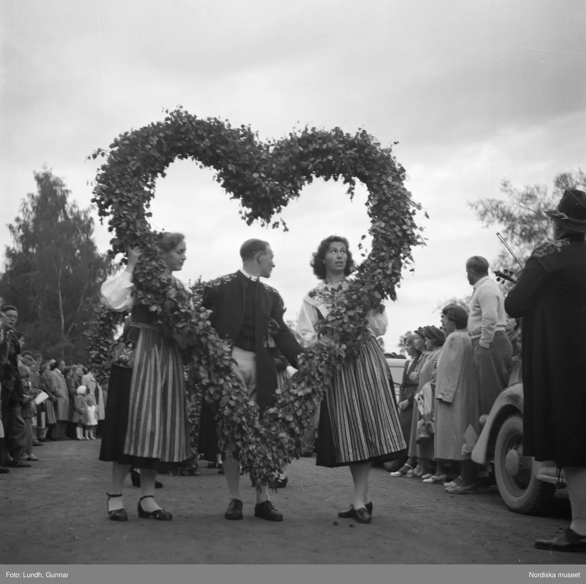 Motiv: (ingen anteckning) ;
Två kvinnor och en man i folkdräkt bär ett lövat hjärta, män i folkdräkt spelar fiol, en folksamling tittar på ett midsommartåg genom ett samhälle, en folksamling "S????dal Leksand".