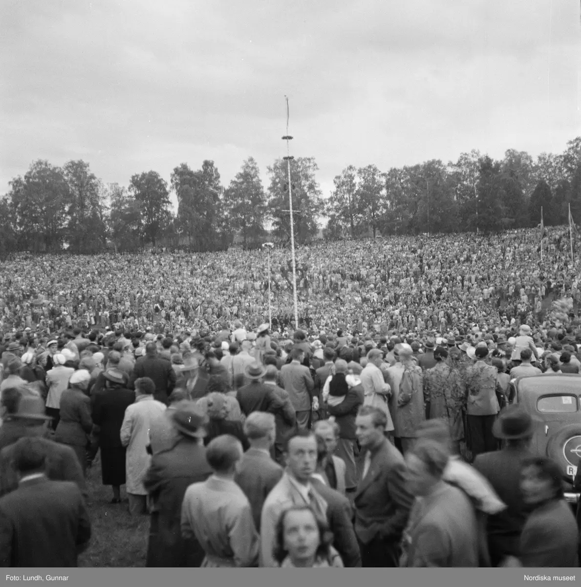 Motiv: (ingen anteckning) ;
En folksamling vid midsommarfirande, en folksamling står på stranden vid en sjö, kvinnor och män i folkdräkt ror en kyrkbåt.