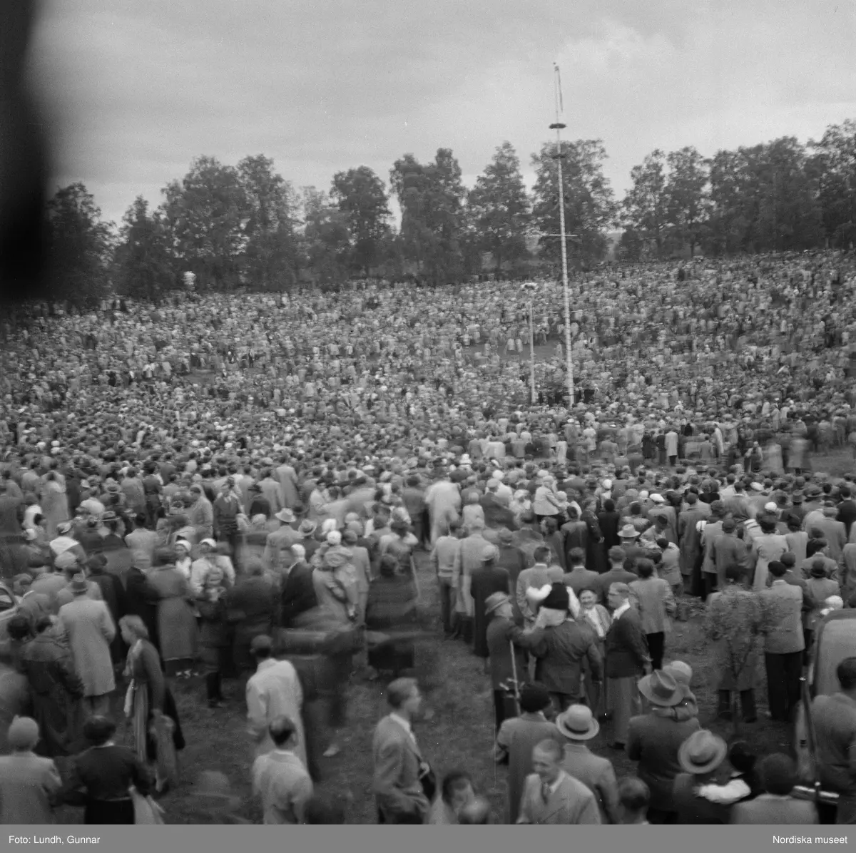 Motiv: (ingen anteckning) ;
En folksamling vid midsommarfirande, en folksamling står på stranden vid en sjö, kvinnor och män i folkdräkt ror en kyrkbåt.