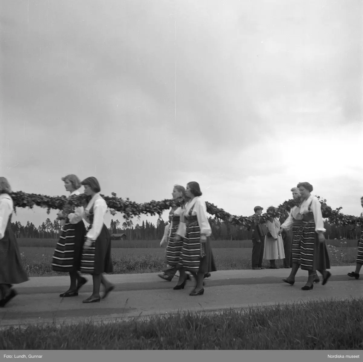 Motiv: (ingen anteckning) ;
Porträtt av en kvinna i folkdräkt och en man som sitter på marken, män och kvinnor i folkdräkt går i ett proccesionståg med flaggor och kvinnor bär en lövad girlang, människor åker på en hästdragen vagn, en folksamling går i ett processionståg.