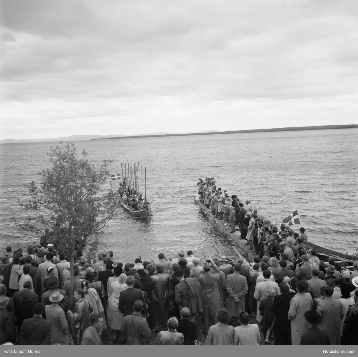 Motiv: (ingen anteckning) ;
En folksamling vid midsommarfirande, en folksamling står på stranden vid en sjö, kvinnor och män i folkdräkt ror en kyrkbåt.