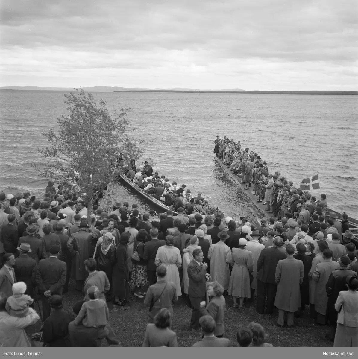 Motiv: (ingen anteckning) ;
En folksamling vid midsommarfirande, en folksamling står på stranden vid en sjö, kvinnor och män i folkdräkt ror en kyrkbåt.