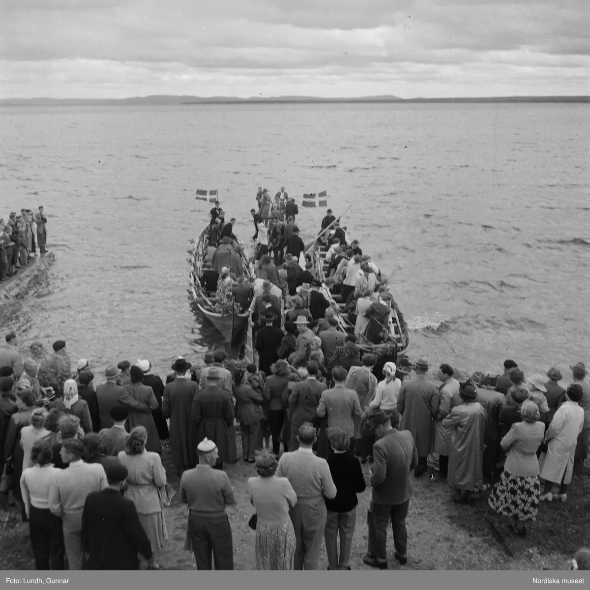 Motiv: (ingen anteckning) ;
En folksamling står vid en brygga med två kyrkbåtar, kvinnor och män ror kyrkbåtar i samband med midsommarfirande.