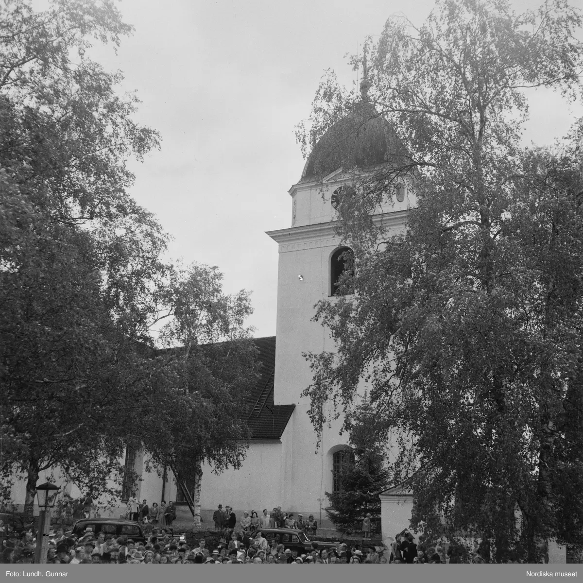 Motiv: (ingen anteckning) ;
En folksamling vid Rättviks kyrka, en folksamling står vid en brygga när kvinnor och män går ombord på två kyrkbåtar.