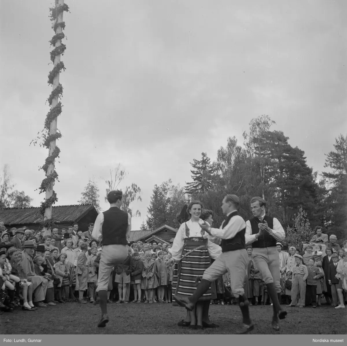 Motiv: (ingen anteckning) ;
Kvinnor och män i folkdräkt dansar vid en midsommarstång inför en publik vid midsommarfirande, tre hästskulpturer 2Jemt-Olofs hästar" (träsnidaren Jemt Olov Persson), porträtt av en man som står vid en husvägg, porträtt av en kvinna som står på en balkong.