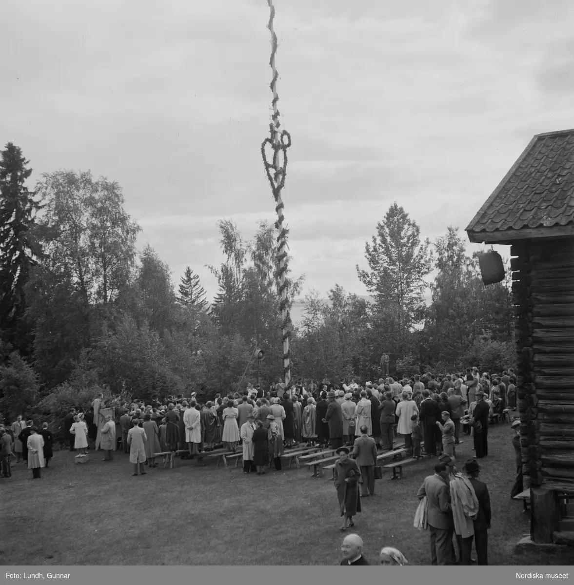 Motiv: (ingen anteckning) ;
Kvinnor och män i folkdräkt dansar vid en midsommarstång inför en publik vid midsommarfirande, tre hästskulpturer 2Jemt-Olofs hästar" (träsnidaren Jemt Olov Persson), porträtt av en man som står vid en husvägg, porträtt av en kvinna som står på en balkong.