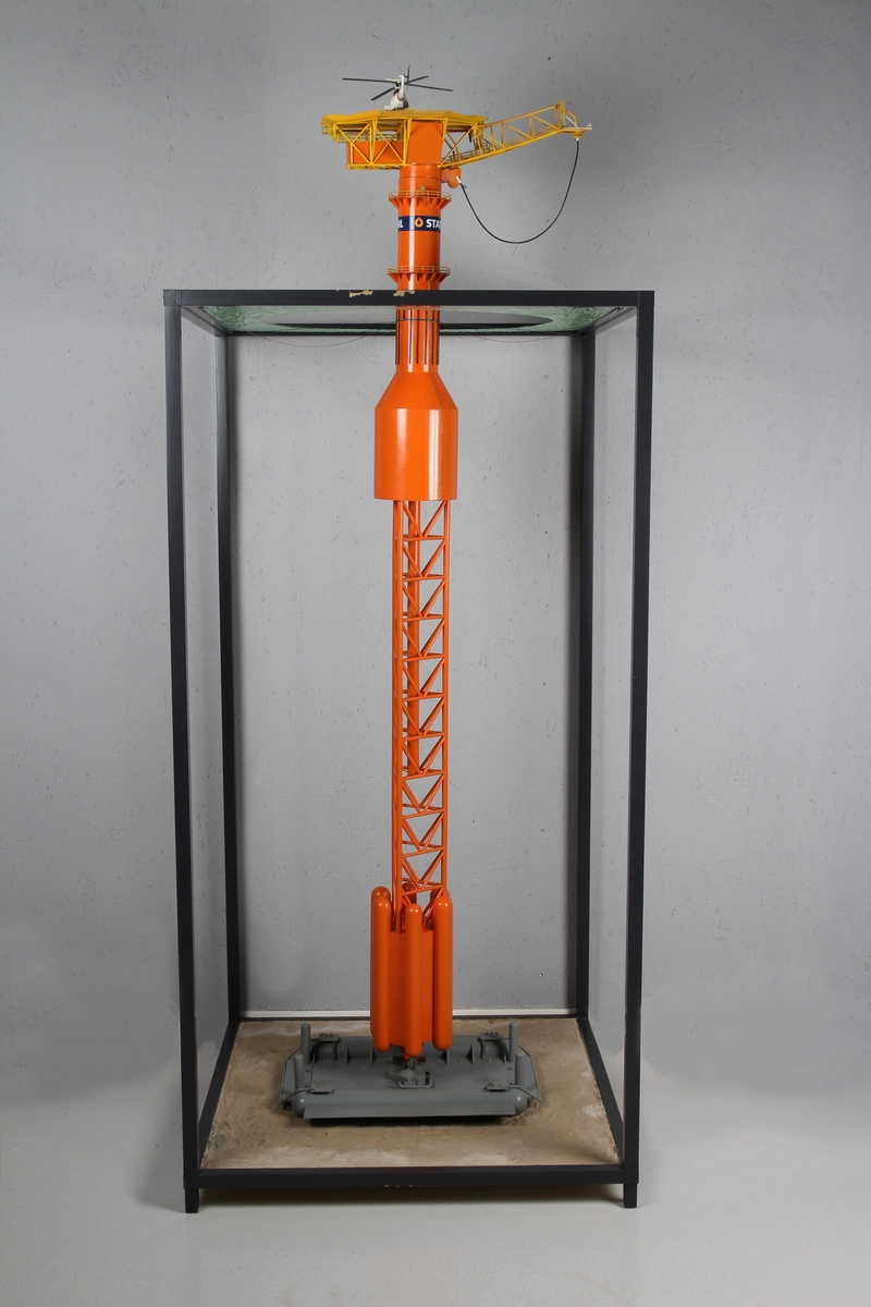 Modell av lastebøyen knyttet til Statfjord A. Malt orange og gul med helikopter på helidekket. Modellen holdes på plass av en aluminiumsramme der bunn og topp forestiller hhv. havbunn og hav.